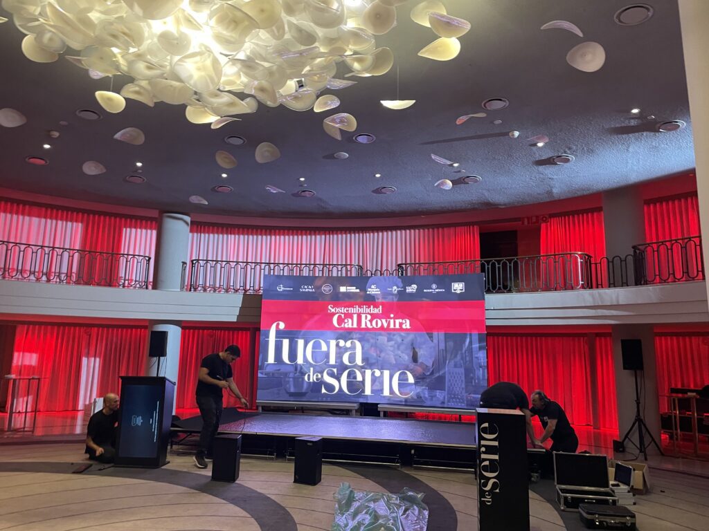 Montaje de evento en salón interior con cortinas rojas, lámparas decorativas en el techo y pantalla de proyección con el texto 'fuera de serie' y logotipos de patrocinadores, trabajadores preparando el equipo de sonido.